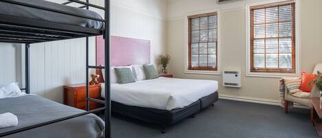A hotel room with bunk beds, a single bed, a chair, a small table, and a window with blinds.