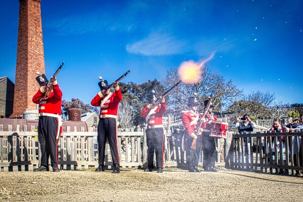 Un grupo de personas uniformadas con atuendo rojo y negro, formadas en fila, con rifles, en un entorno al aire libre con cielo despejado y una estructura de ladrillo al fondo.