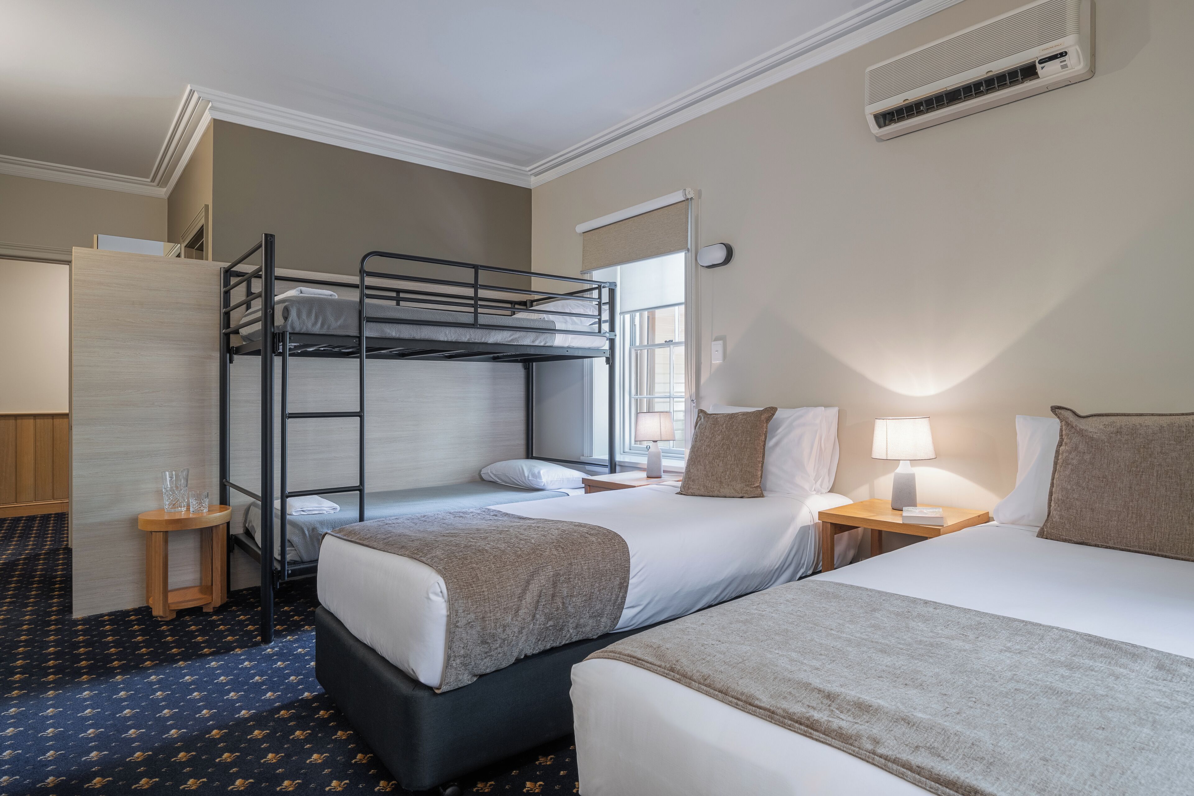 A hotel room with a bunk bed, a single bed, a bedside table with a lamp, and a window with a view.