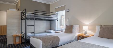 A hotel room with a bunk bed, a single bed, a bedside table with a lamp, and a window with a view.
