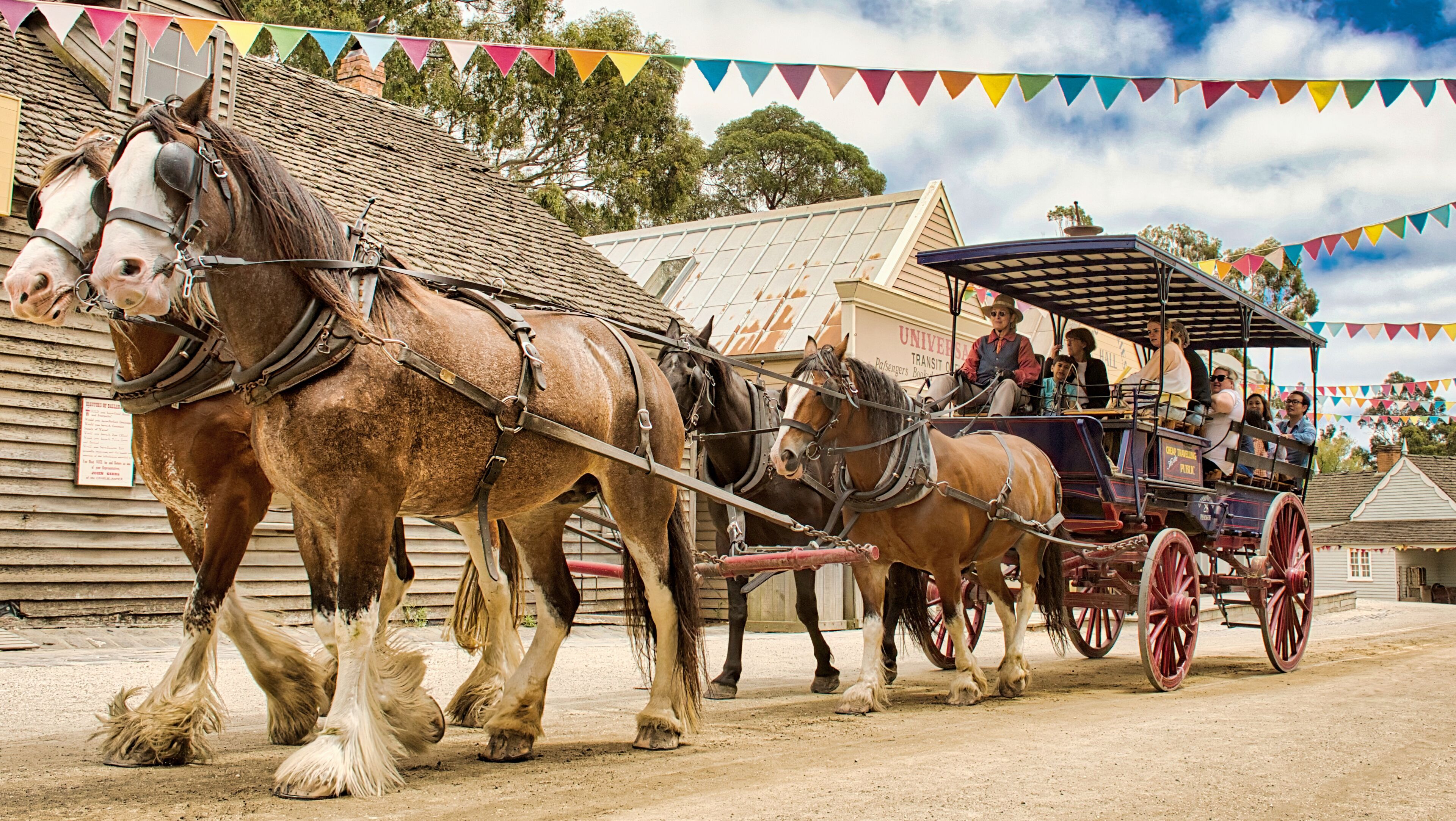 A horse-drawn carriage with passengers, pulled by two large horses, on a dirt road with buildings in the background.