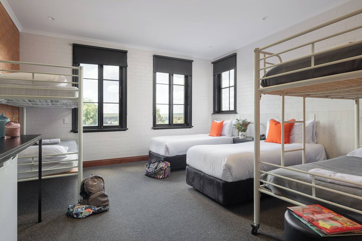 Habitación tipo dormitorio con literas, un escritorio y una silla. Hay dos ventanas con persianas negras, una pared de ladrillo y una alfombra gris.