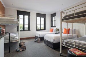 Habitación tipo dormitorio con literas, un escritorio y una silla. Hay dos ventanas con persianas negras, una pared de ladrillo y una alfombra gris.