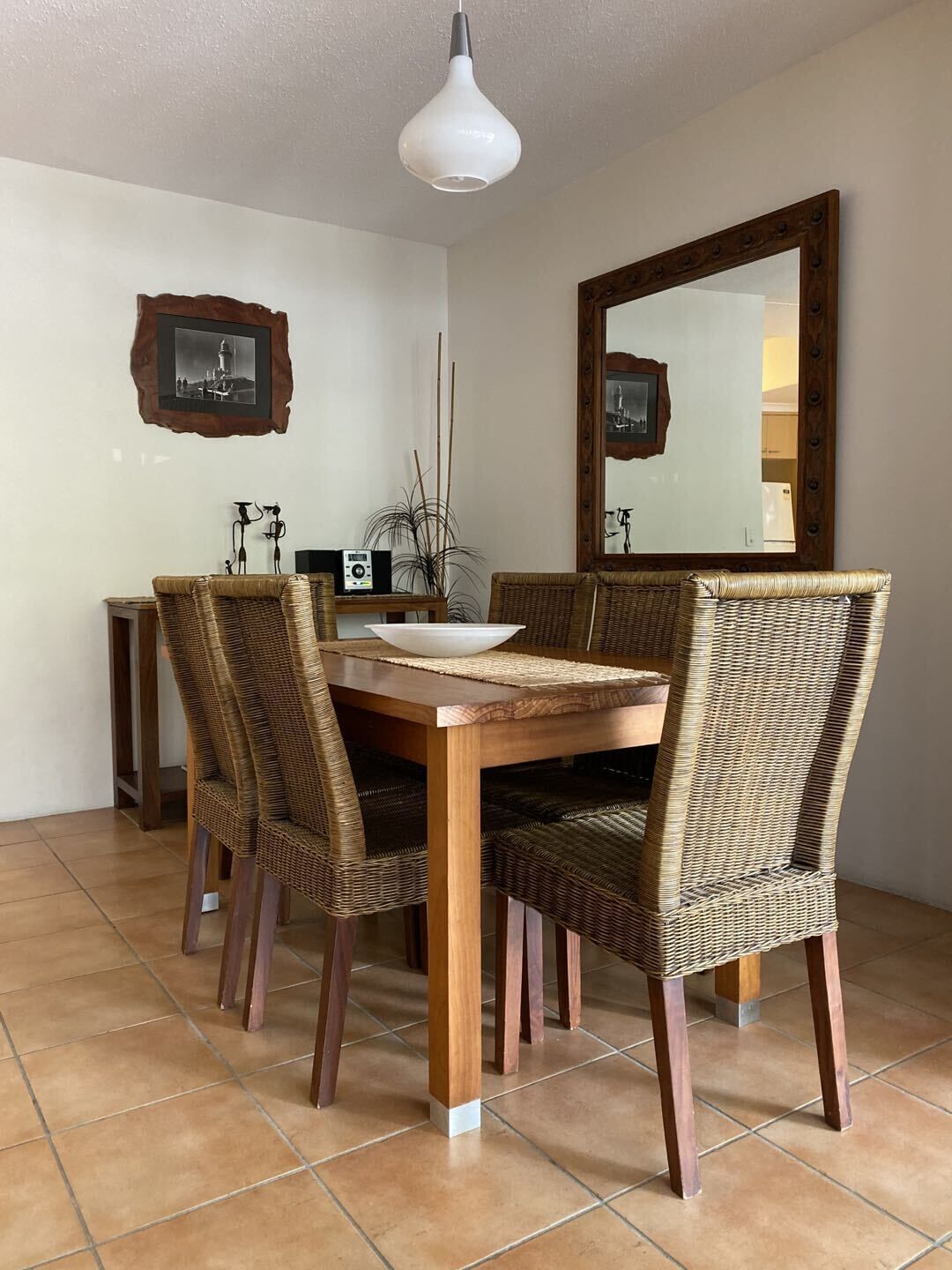 A dining area with a wooden table, wicker chairs, a white pendant light, and a large mirror on the wall.
