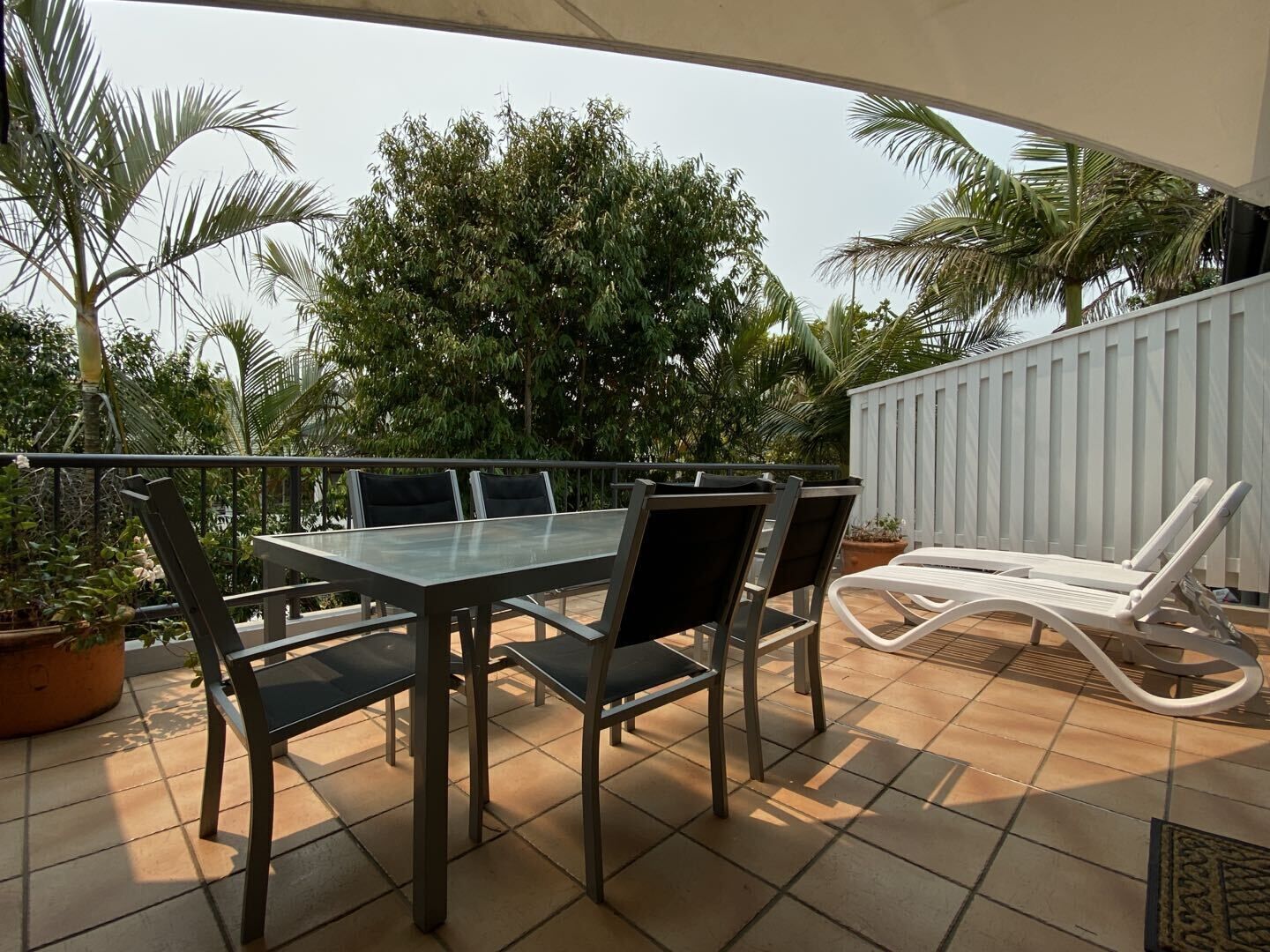 An outdoor patio with a glass-top table and chairs, surrounded by potted plants and a white fence.