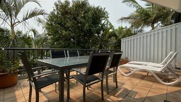An outdoor patio with a glass-top table and chairs, surrounded by potted plants and a white fence.