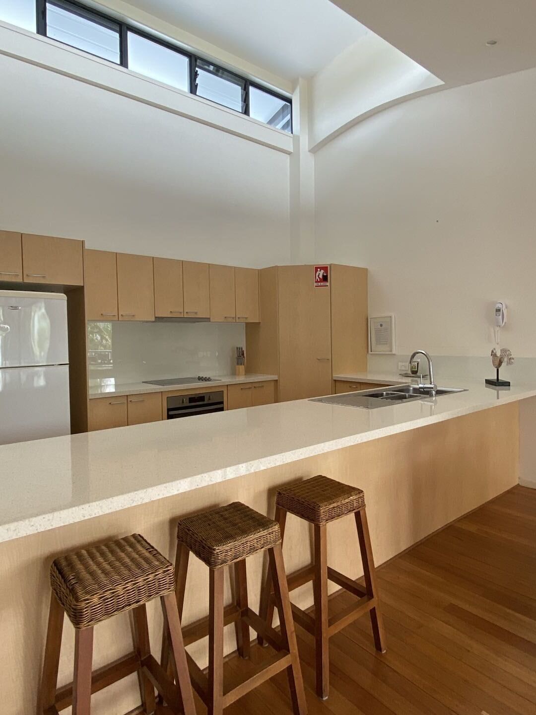A modern kitchen with wooden cabinets, a white countertop, and a built-in dishwasher. There are three wicker stools at the counter.