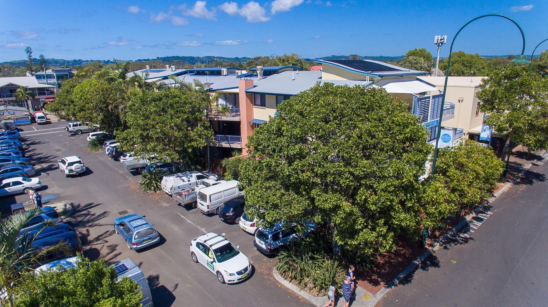 A parking lot with several cars and a few delivery trucks. There are buildings with solar panels on the roofs and a large tree in the foreground.