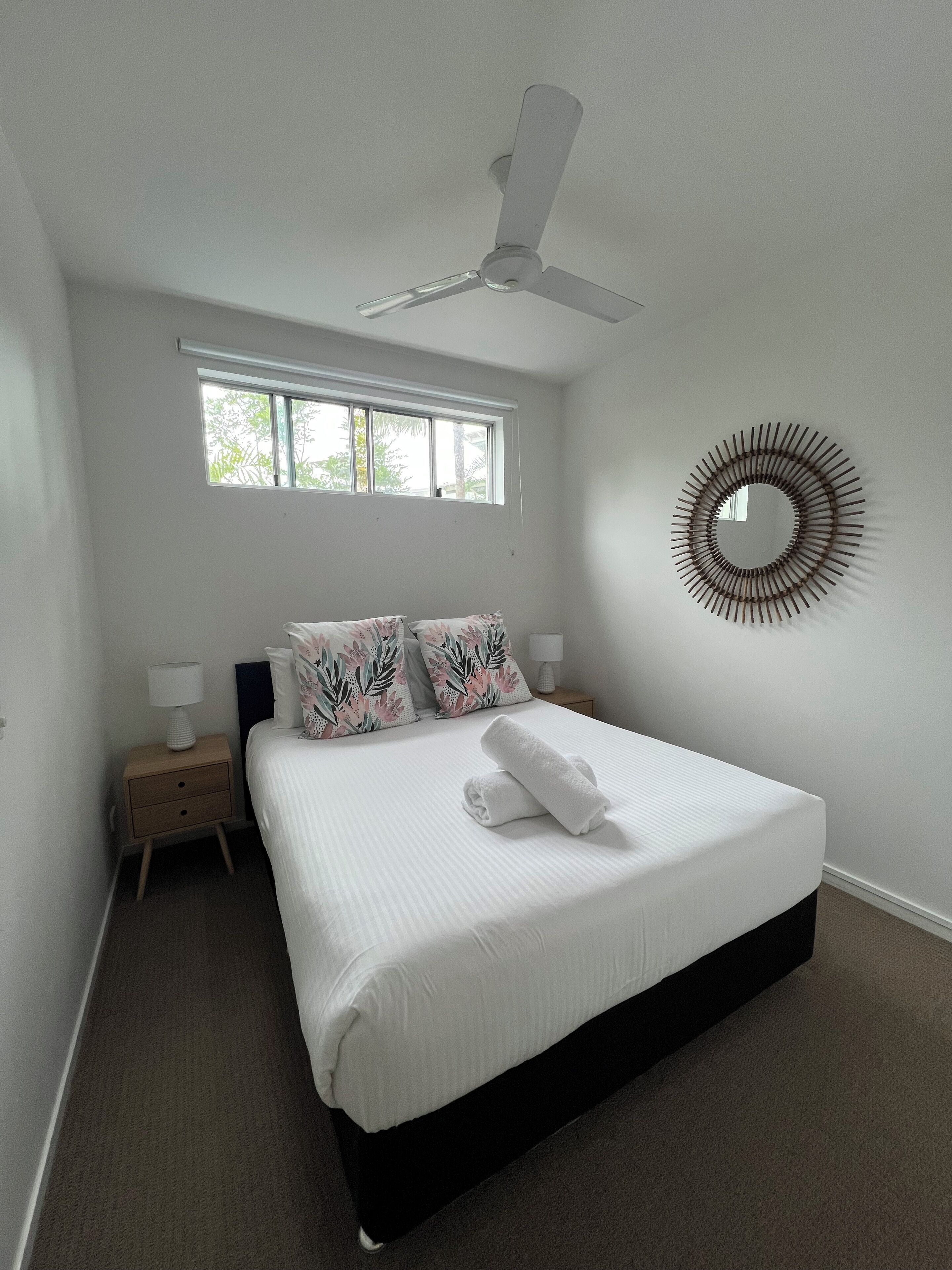 Ein Schlafzimmer mit einem Bett, einem Deckenventilator, einem Fenster, einem Nachttisch und einem an der Wand befestigten Spiegel.
