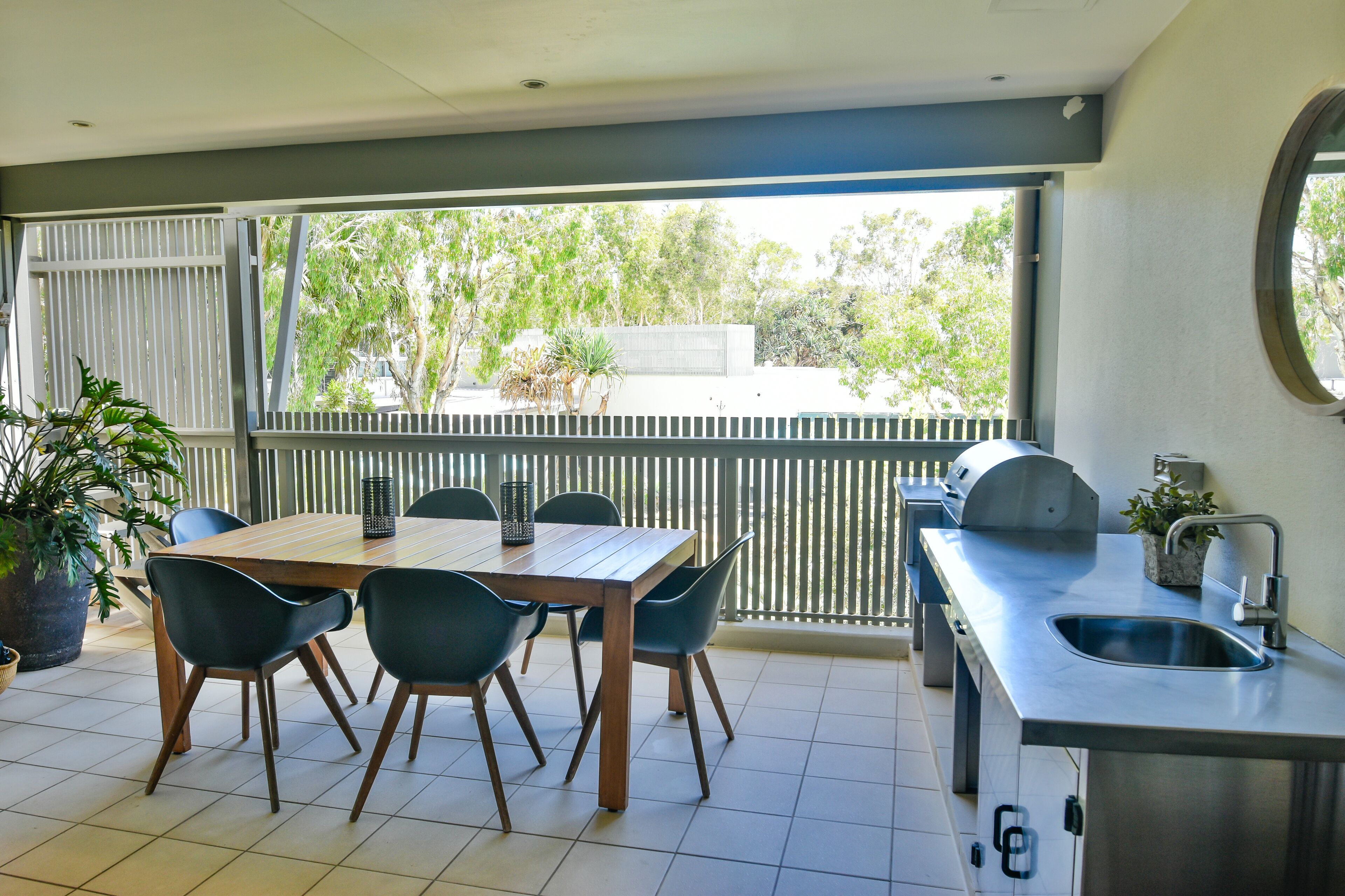A dining area with a wooden table and chairs, a kitchen with a sink, and a view of trees through large windows.