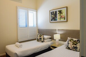 Habitación de hotel con dos camas, una mesita de noche con lámpara, un cuadro enmarcado de palmeras y una ventana con persianas.