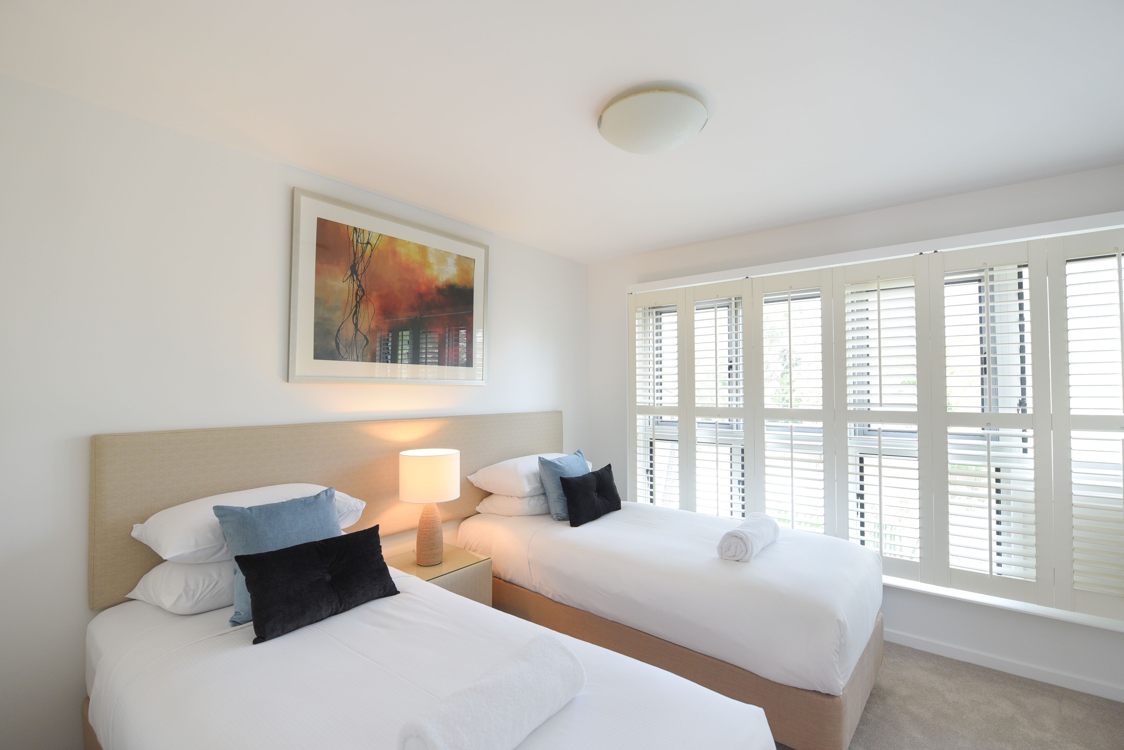Une chambre d’hôtel avec deux lits, un tableau au mur, une fenêtre avec des stores et une lampe de chevet.