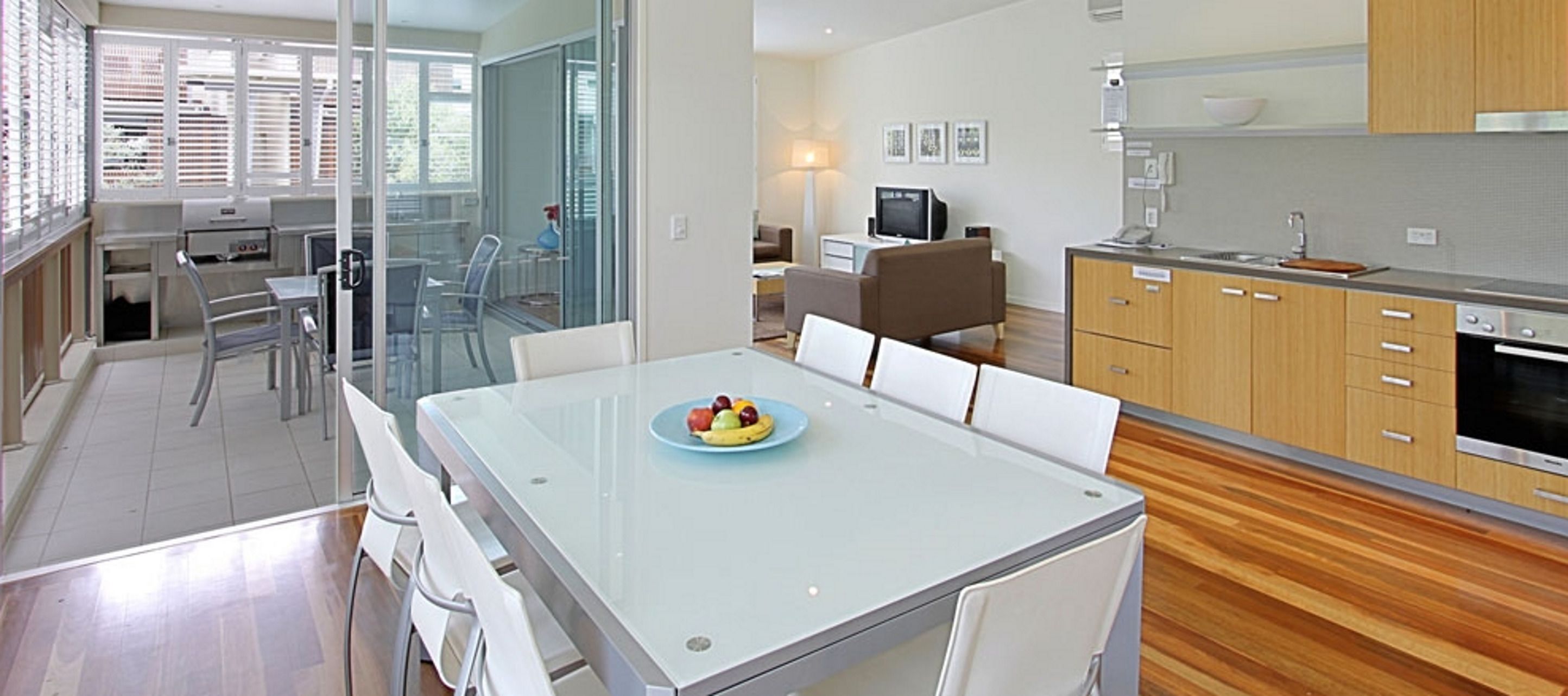Una cocina moderna con comedor, que incluye una mesa de vidrio con frutero, armarios de madera y un televisor al fondo.