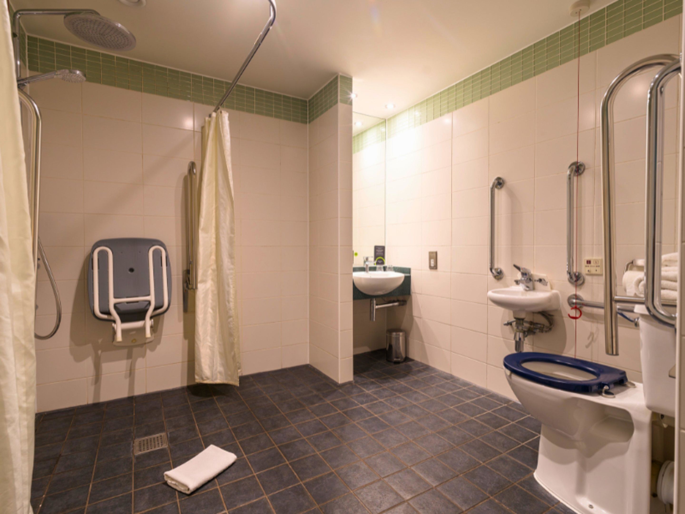 Une salle de bain équipée d’une douche, de toilettes avec un siège surélevé, d’un lavabo et d’une barre d’appui.