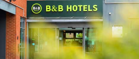 A B&B hotel entrance with a glass facade, a brick building to the left, and a sign above the door.