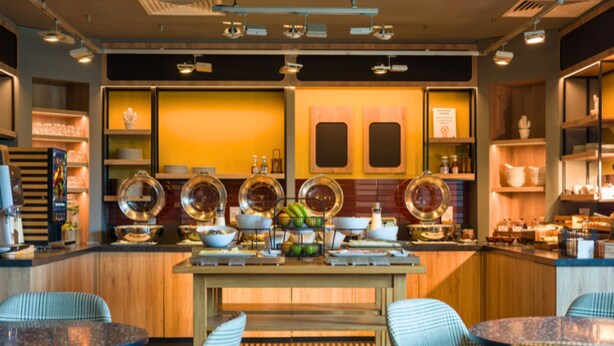 A modern kitchen with wooden cabinets, a central island with seating, and various kitchenware displayed on shelves. - B&B HOTEL Ipswich (Ipswich)