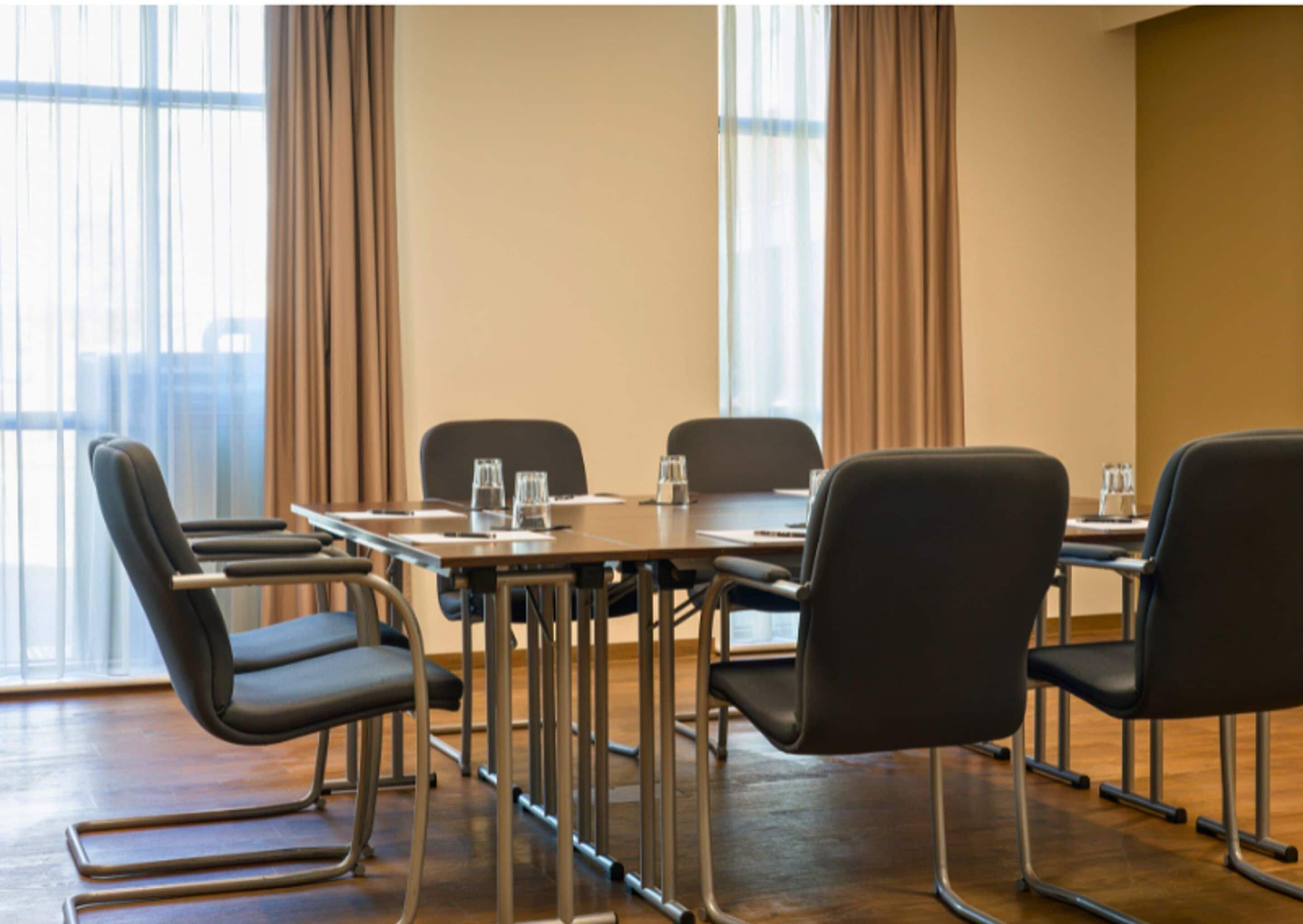 A conference room with a long table, four chairs, and a window with curtains.