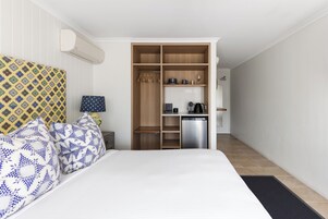 Un dormitorio con cama, almohadas estampadas, una mesita de noche, una lámpara, un armario y una pequeña cocina americana.