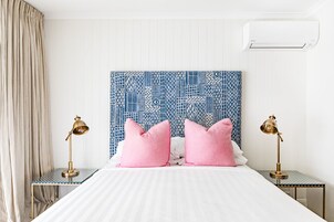 Una cama bien hecha con ropa blanca y dos almohadas rosas, flanqueada por dos mesitas de noche con lámparas doradas.