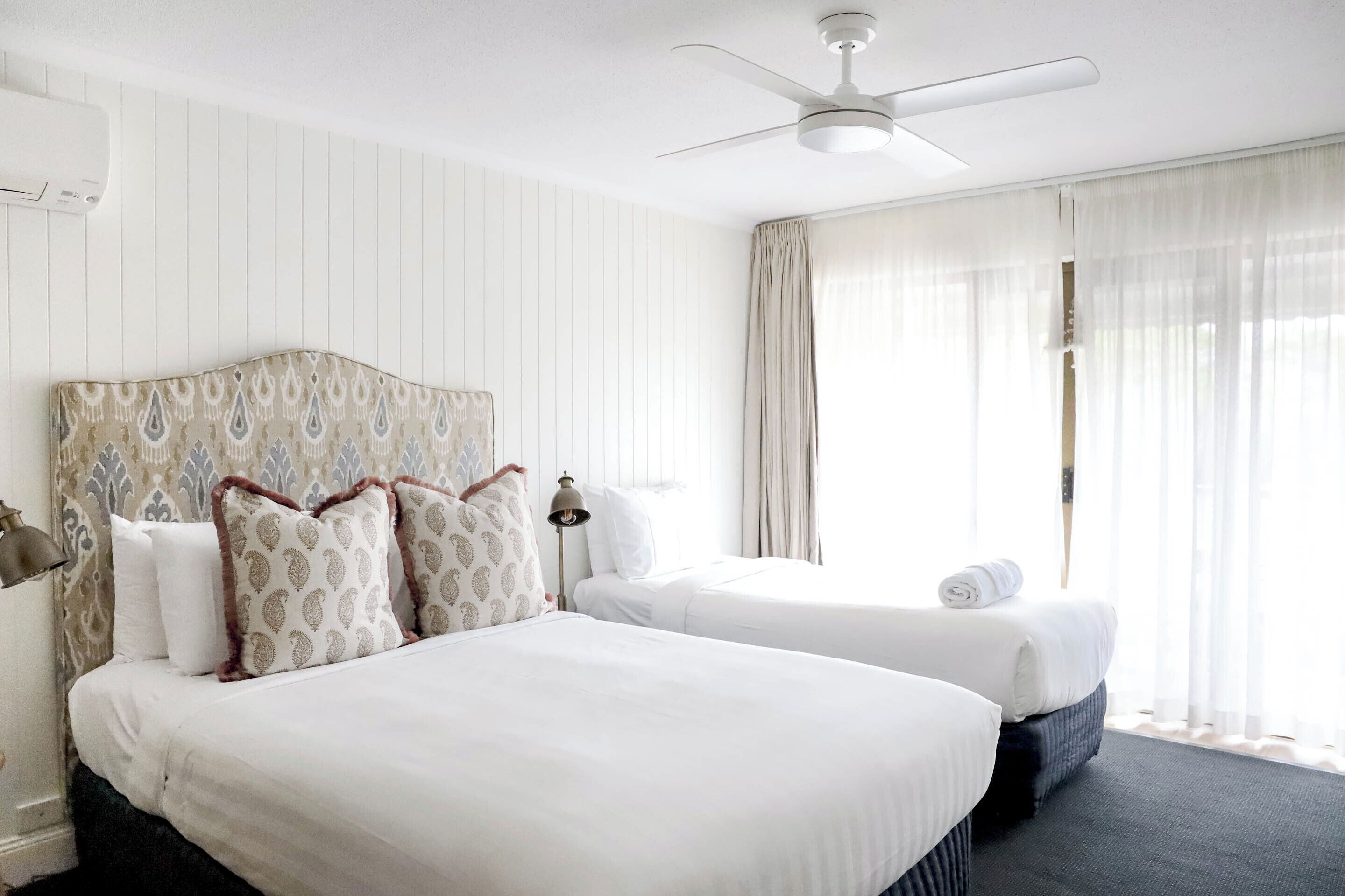 A hotel room with two beds, a ceiling fan, and a large window with sheer curtains.