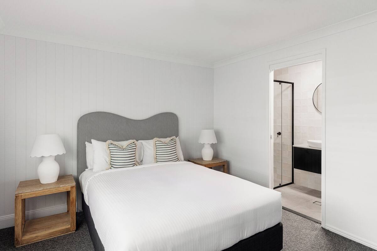 Ein modernes Hotelzimmer mit einem großen Bett, zwei Nachttischen mit Lampen, einem Holzkopfende und einem durch eine offene Tür einsehbaren Badezimmer.