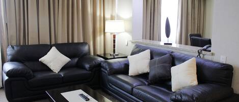 Quarto de hotel com sofá de couro preto, uma mesinha com controle remoto e uma luminária.