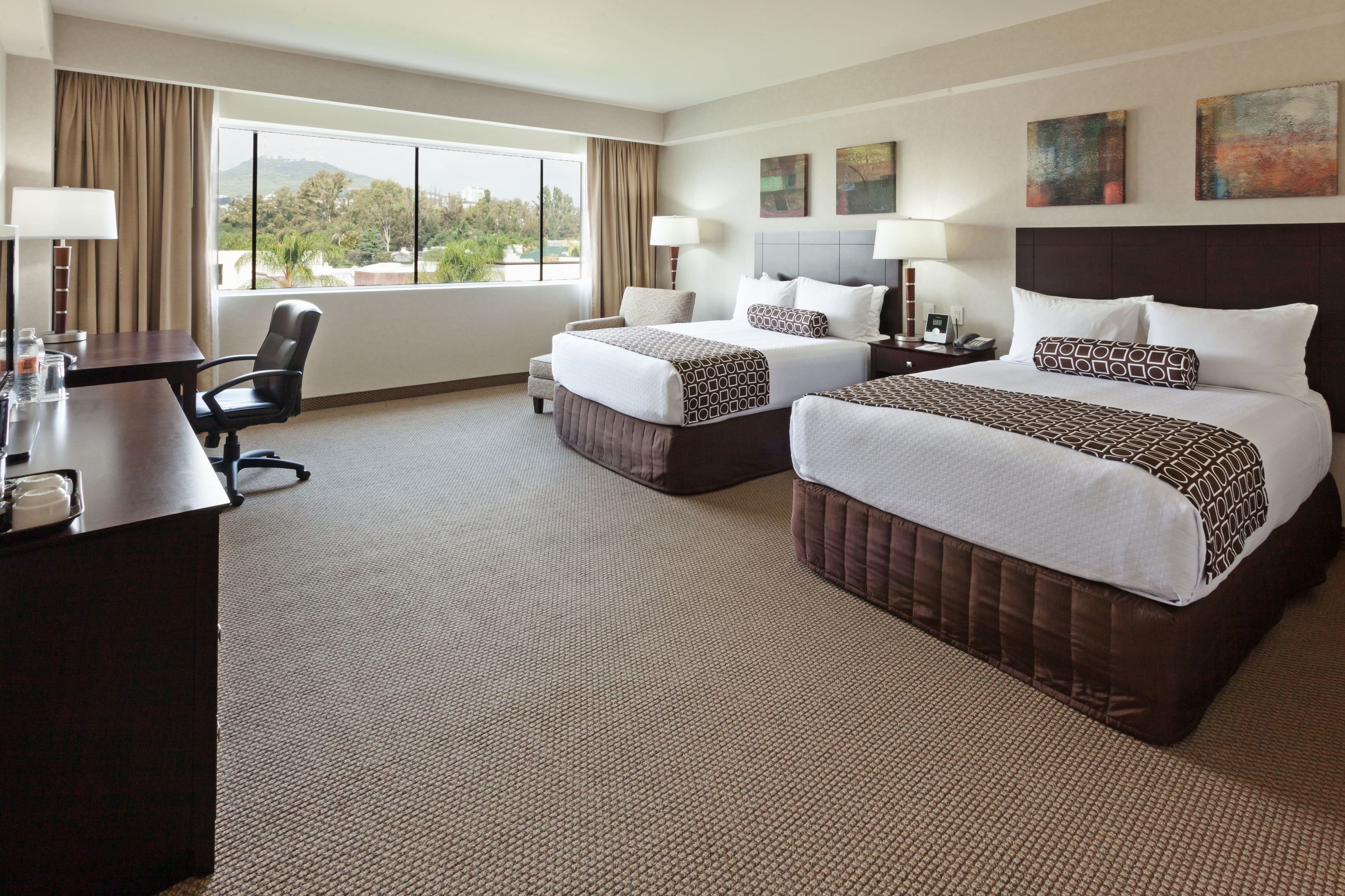 A hotel room with two beds, a desk, a chair, and a large window offering a view of greenery.