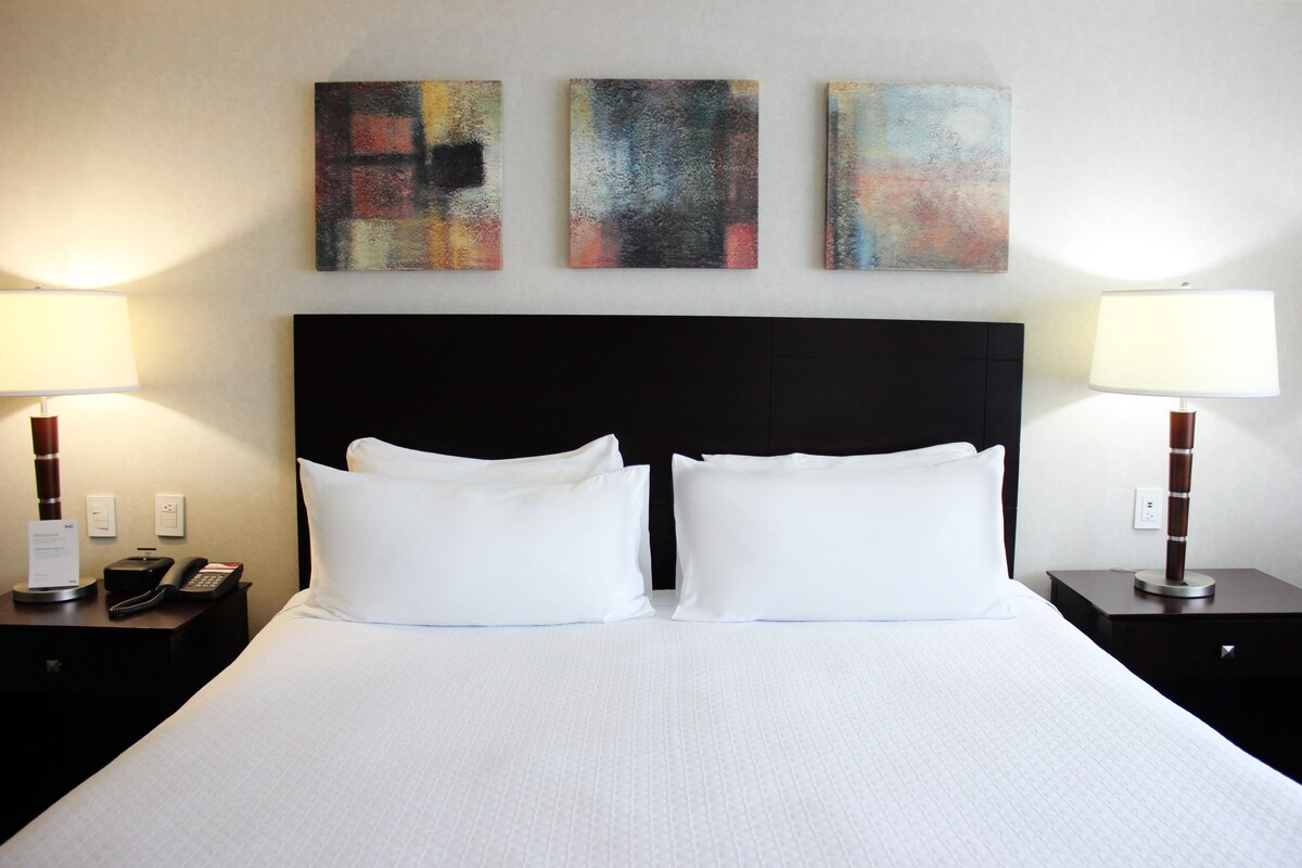A hotel room with a large bed, two bedside tables, a black headboard, and three abstract paintings on the wall.