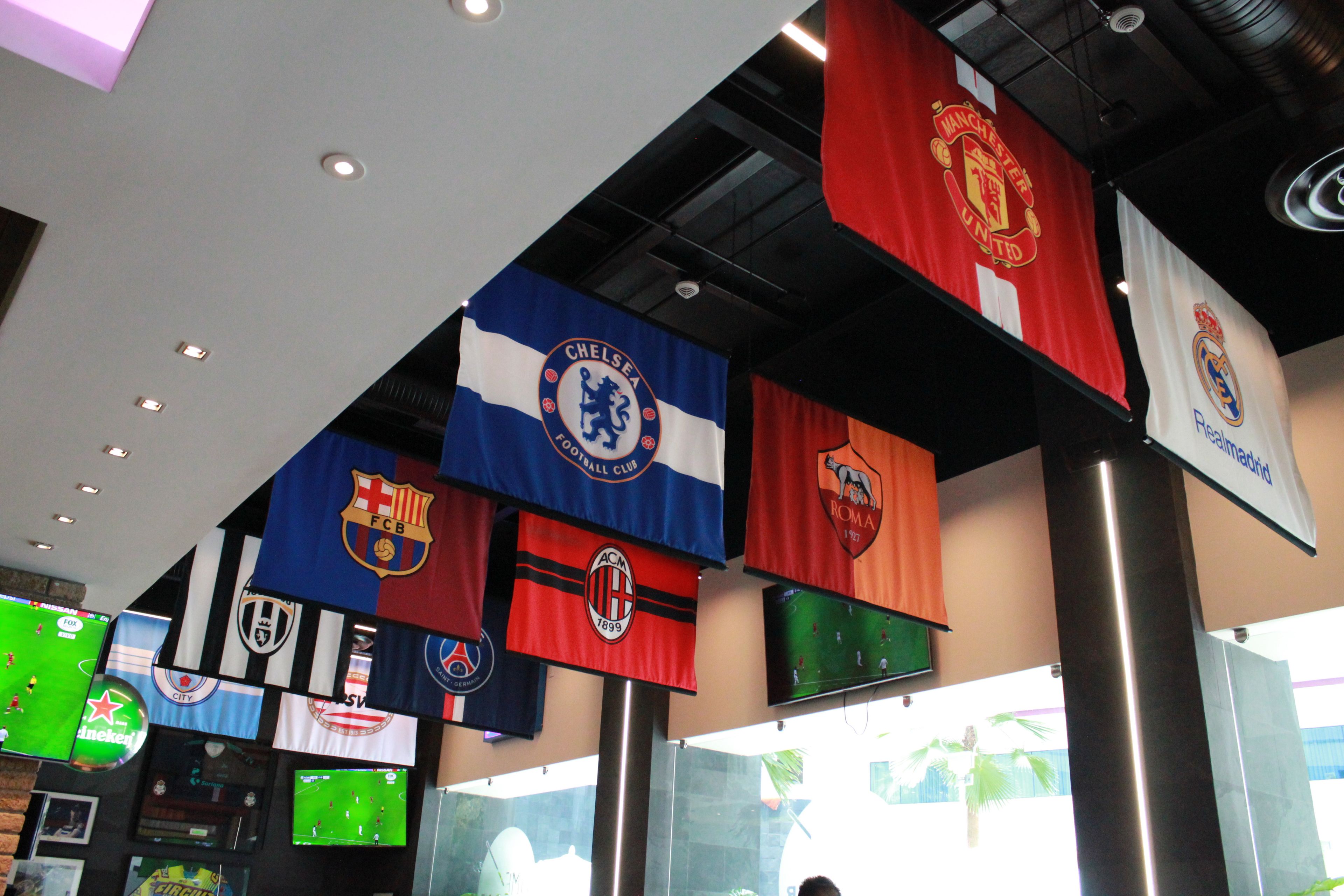 Un bar sportif doté de plusieurs écrans diffusant des matchs de football, et dont le plafond est décoré de drapeaux représentant différents clubs de football.