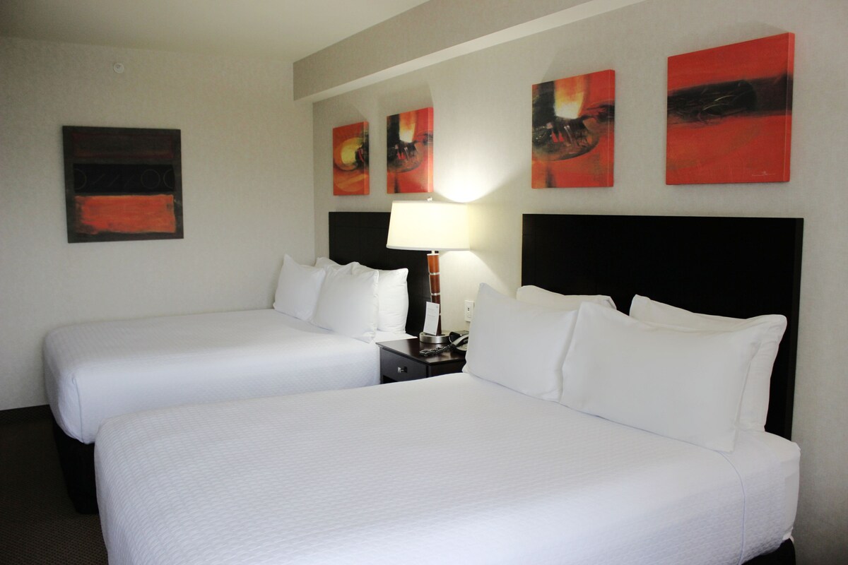 A hotel room with two beds, a nightstand with a lamp, and four abstract paintings on the wall.