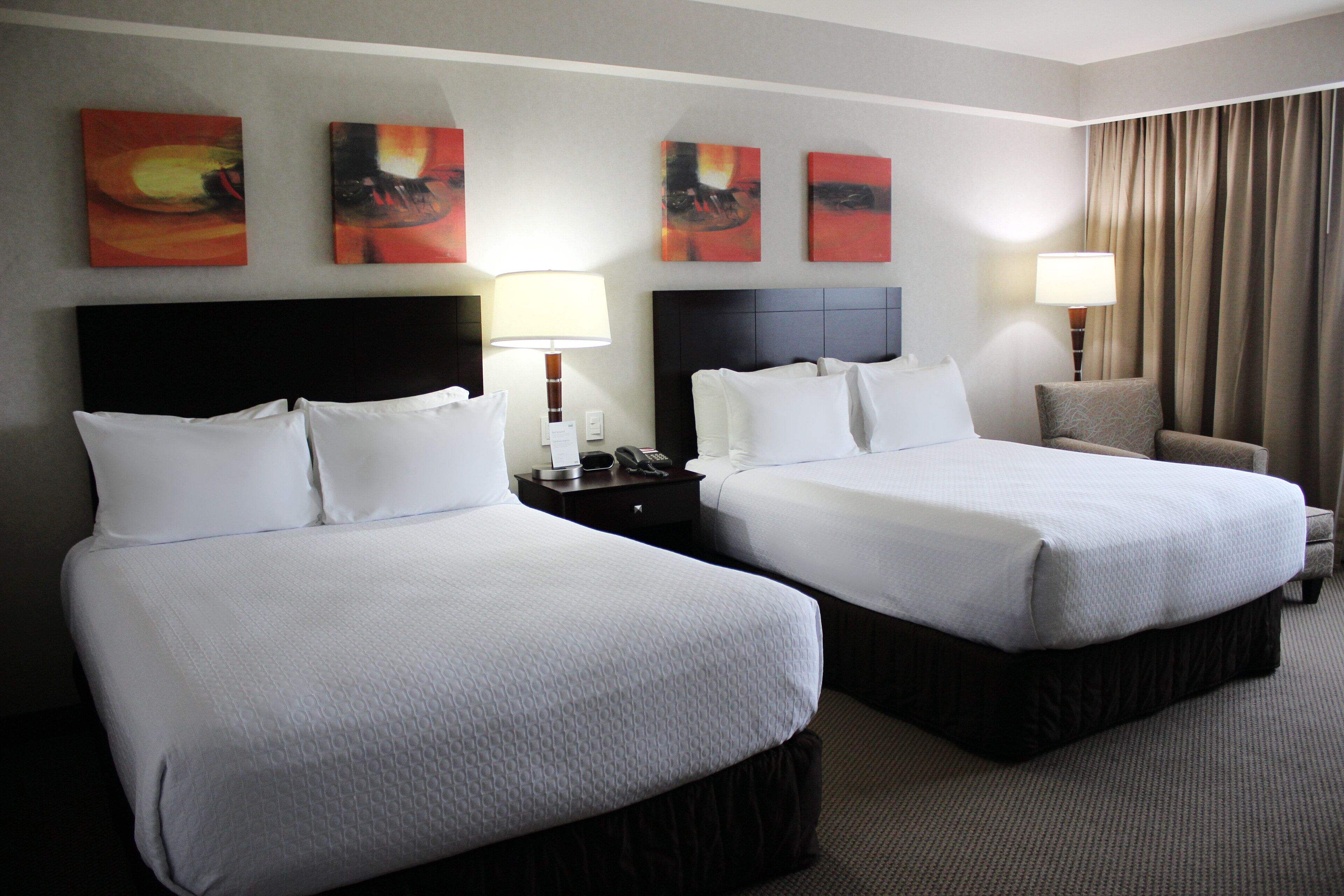 A hotel room with two beds, a nightstand with a lamp, a chair, and artwork on the wall.