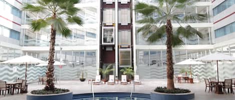 Una piscina interior moderna con techo de vidrio, rodeada de palmeras y zonas de descanso.