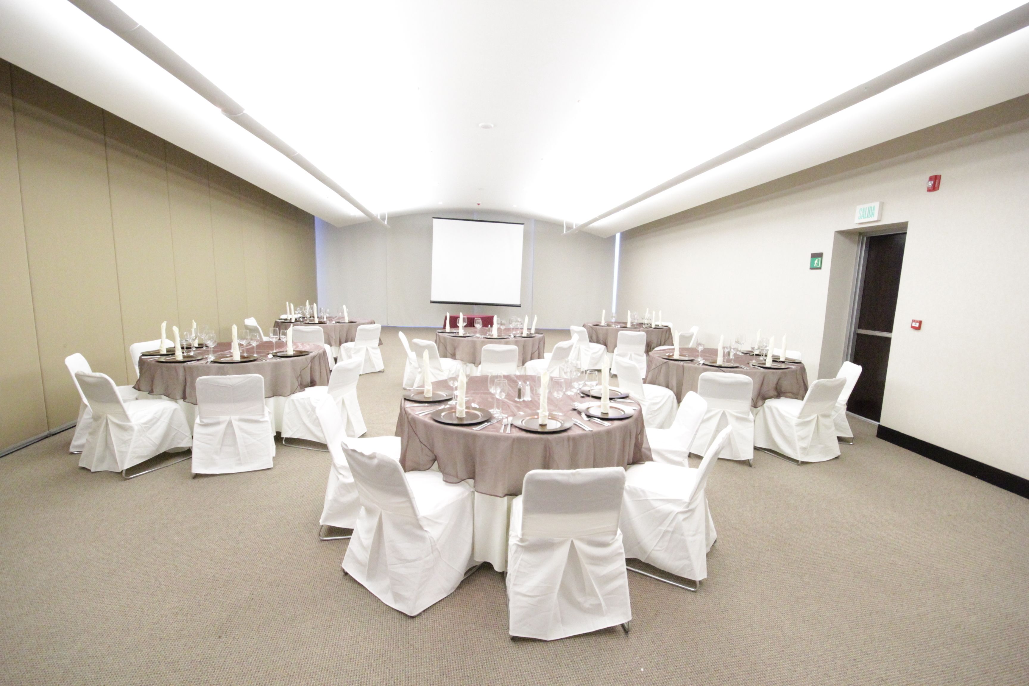Une salle de conférence avec des tables rondes, des chaises blanches et un écran de projection.