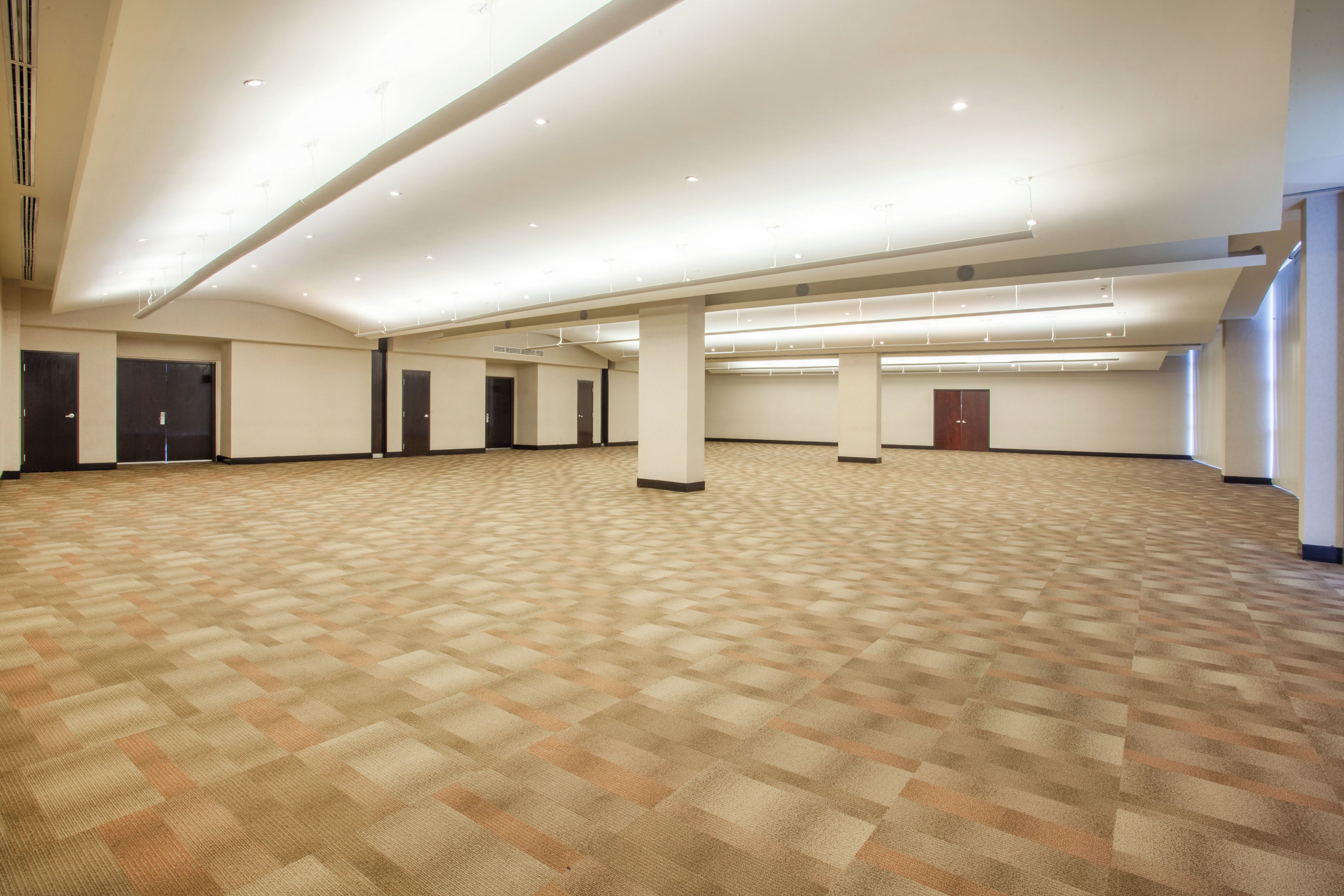 Amplia habitación de hotel vacía con alfombra estampada, varias puertas y un techo alto con iluminación empotrada.