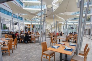 A spacious indoor dining area with multiple tables and chairs, large overhead umbrellas, and a modern architectural design with glass railings. - Crowne Plaza Leon by IHG (León)
