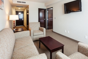 A hotel room with a sofa, armchair, and a small table. There is a TV mounted on the wall and a lamp on a side table. - Crowne Plaza Leon by IHG (León)