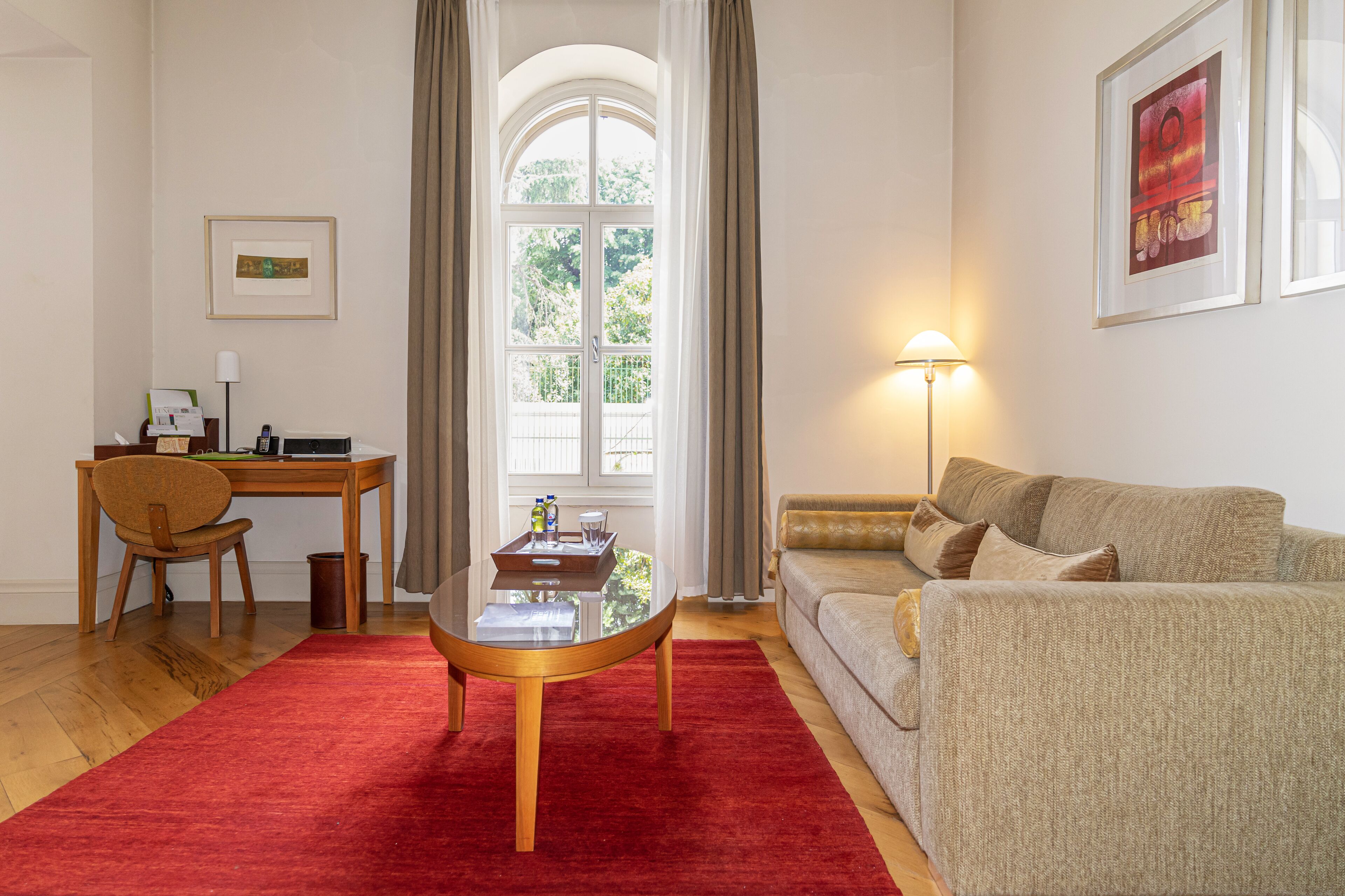 A living room with a red carpet, a wooden coffee table, a beige sofa, a desk with a chair, and a window with curtains.