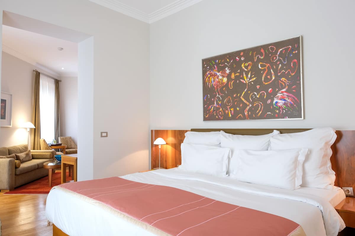 Habitación de hotel con una cama grande, cabecera de madera, un cuadro en la pared, un sofá, una lámpara y una ventana con cortinas.