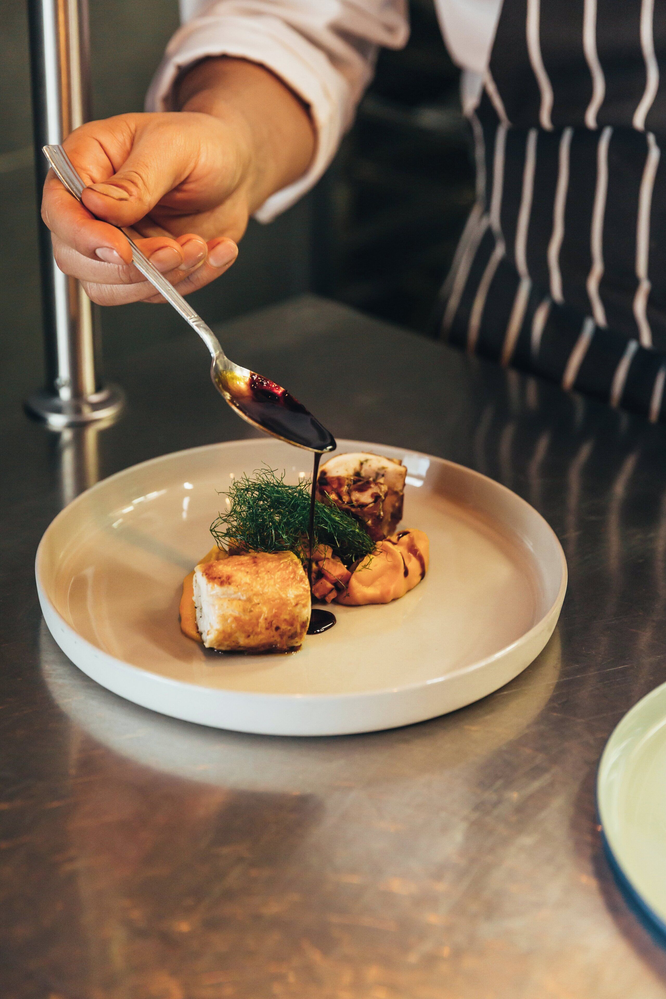 A chef's hand holding a spoon, pouring sauce onto a dish with food items and herbs.