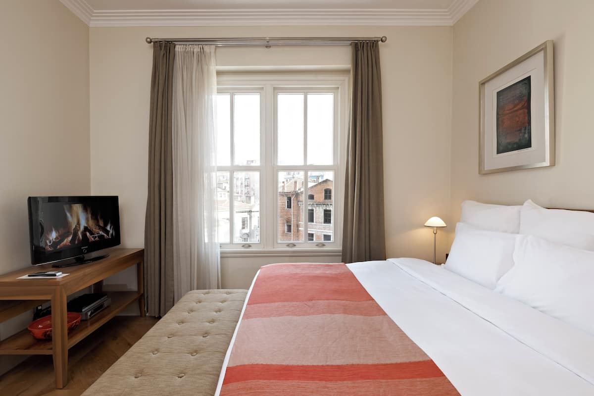 Habitación de hotel con cama, televisor sobre un atril de madera, ventana con cortinas y lámpara.