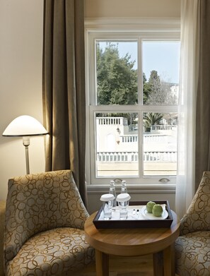 A room with two patterned sofas, a round wooden table with a tray of glasses and bottles, a lamp, and a window with a view of a white fence and trees.