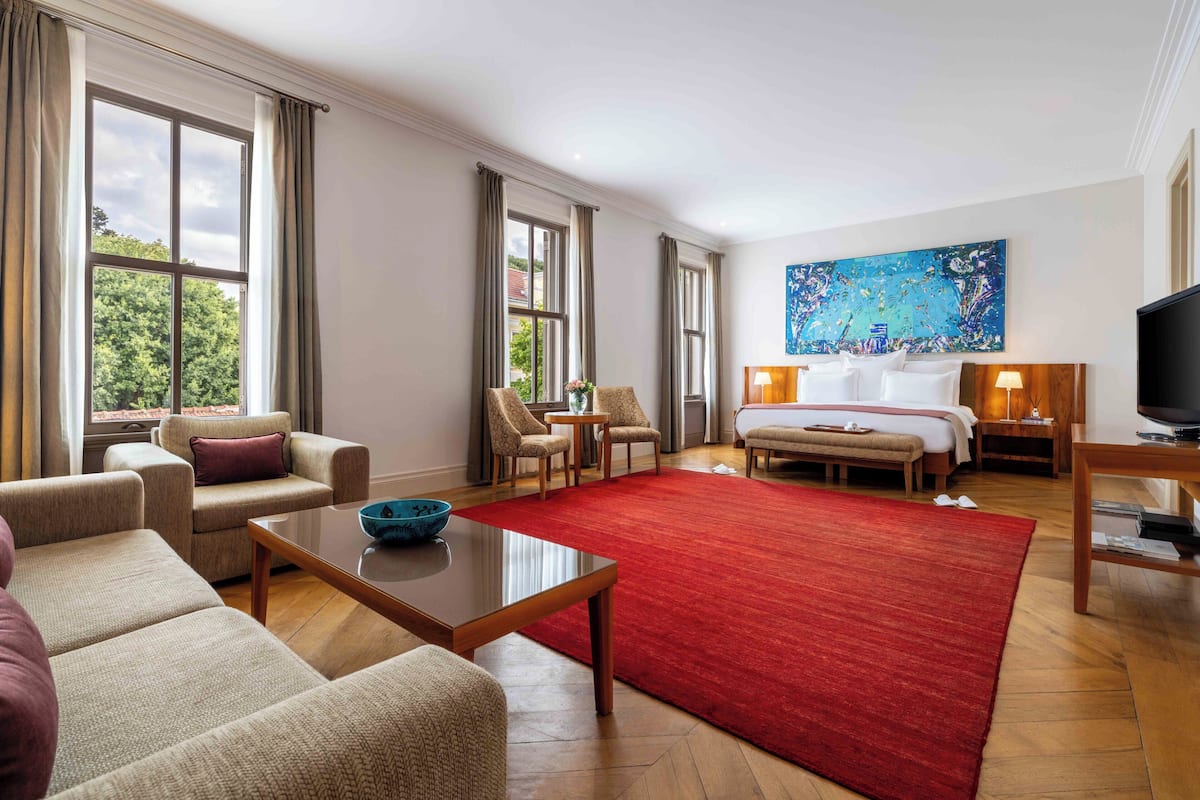 Habitación de hotel con un ventanal grande, alfombra roja, cama, televisión y un cuadro en la pared.