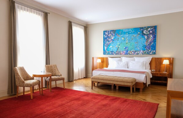 A hotel room with a large bed, two armchairs, a small table, a red rug, and a large abstract painting on the wall.