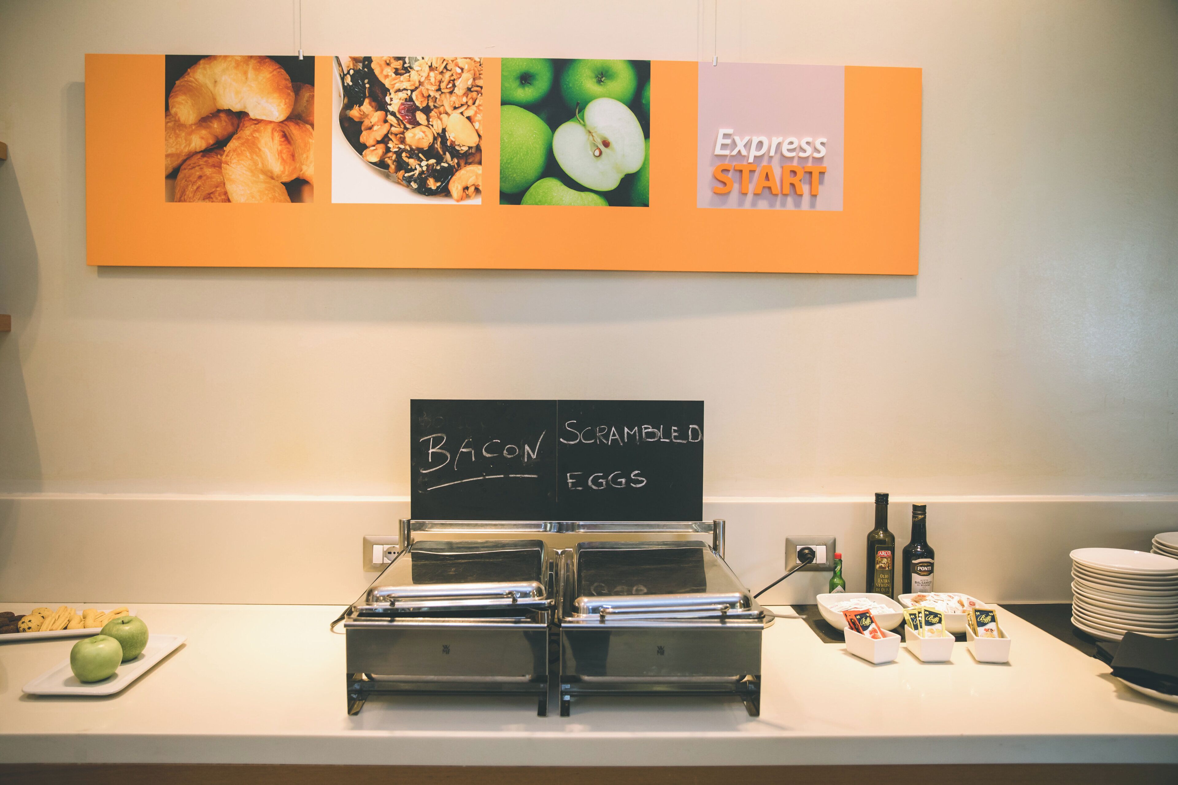 Ein Frühstücksbuffet mit Schildern, die Speck und Rührei anbieten, einer Kaffeestation und einer an der Wand befestigten Speisekarte.