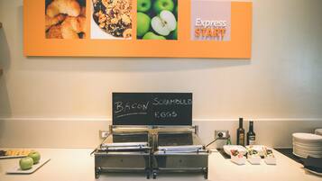 Ein Frühstücksbuffet mit Schildern, die Speck und Rührei anbieten, einer Kaffeestation und einer an der Wand befestigten Speisekarte.