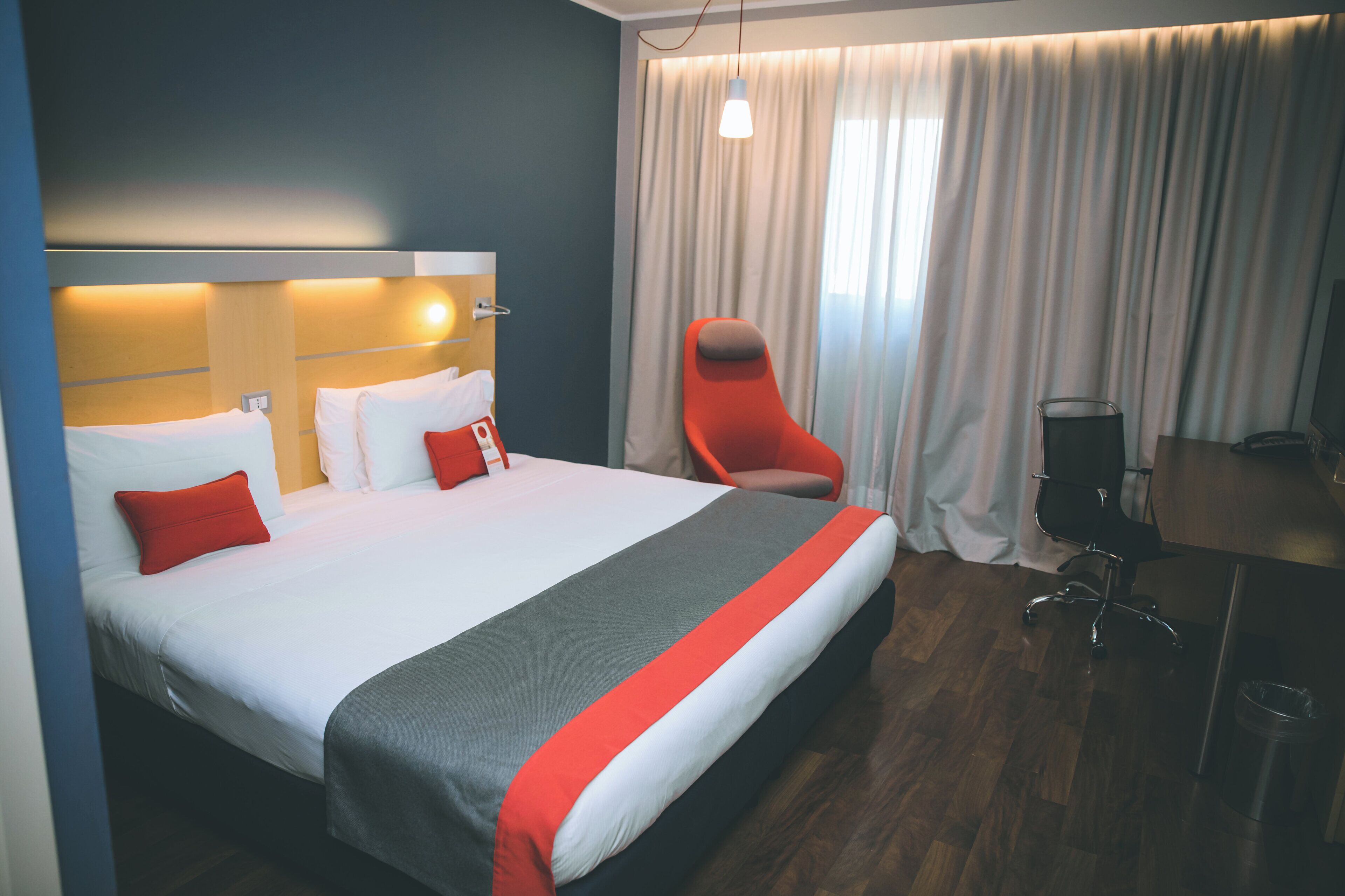 A hotel room with a bed, a chair, a desk, and a window with curtains.