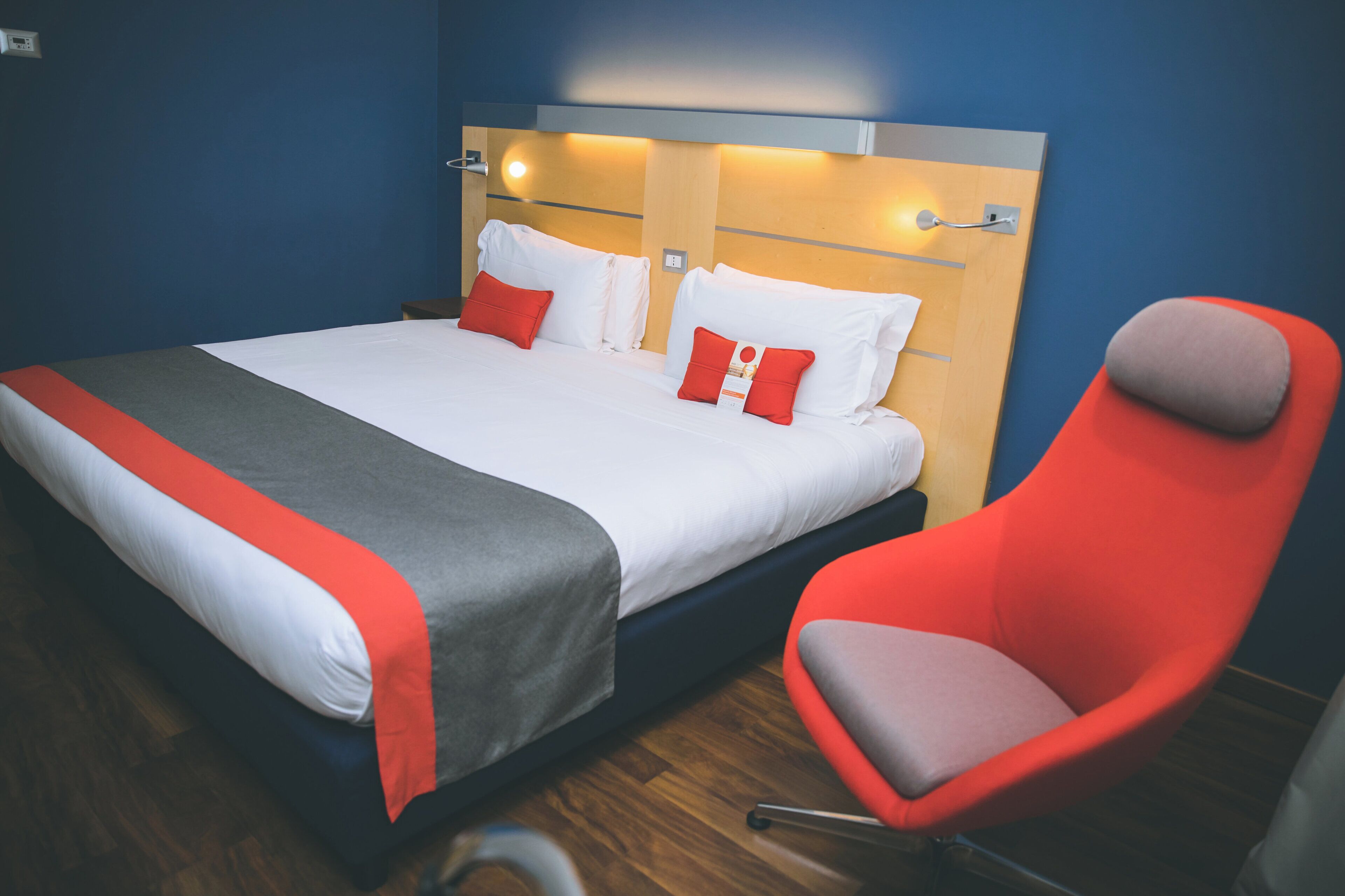 Una habitación de hotel con cama, un sillón rojo y un cabecero de madera con iluminación integrada.