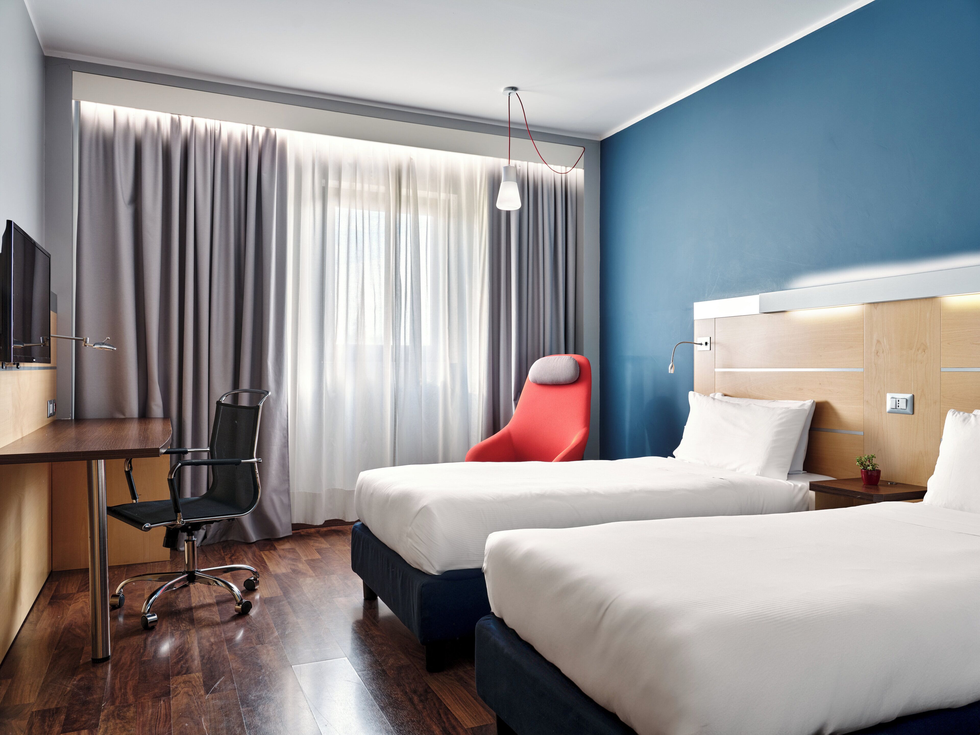 Habitación de hotel con dos camas, un escritorio con silla, televisor y un ventanal con cortinas.