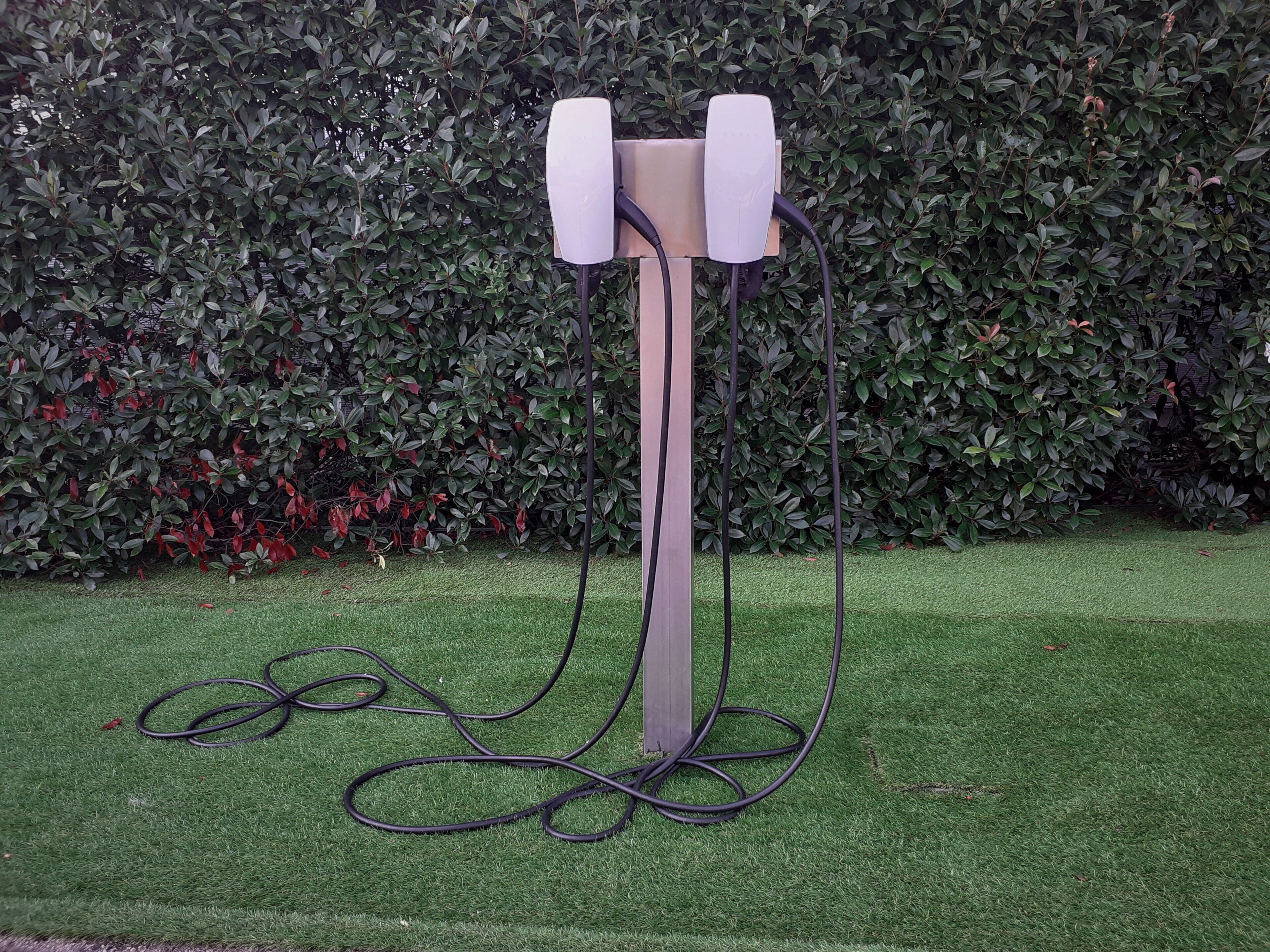 A stand-alone, modern, white, vertical structure with a cable connected to it, set against a backdrop of dense green foliage.