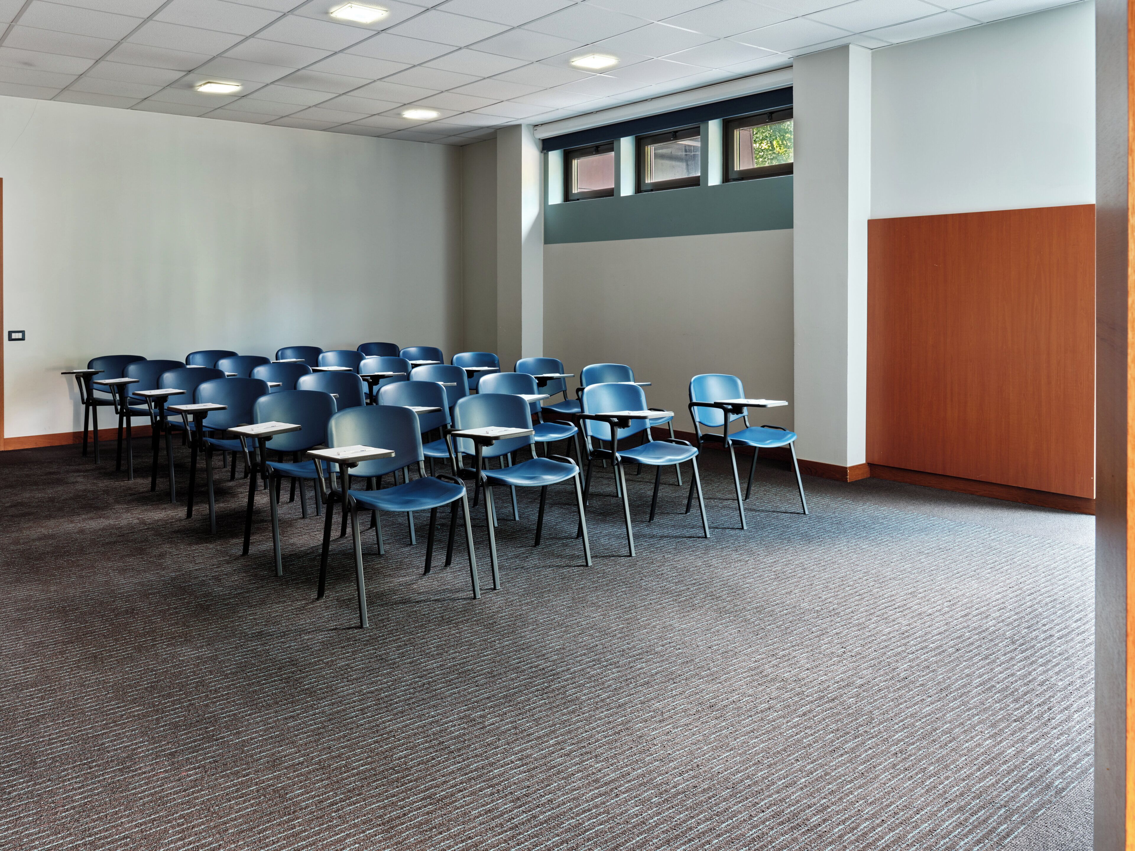 A conference room with rows of chairs and tables, a carpeted floor, and a window with blinds.